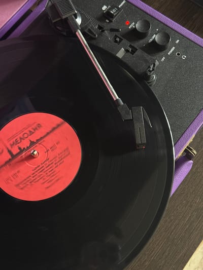 A record player sits on a dark wooden surface. The player is black with a purple trim. A black vinyl record with a red label is on the turntable. The needle is positioned on the record. The player has knobs and switches for power, volume, pitch, and phono/off/on. The label on the vinyl says "MEADLAWS".