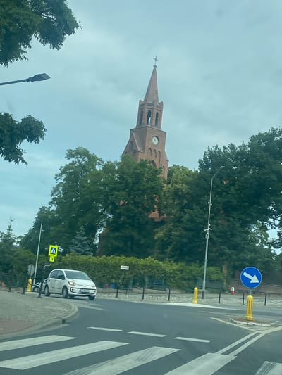 This is a daytime, eye-level, outdoor photograph taken from a vehicle, likely at a crosswalk. The focal point of the image is a red brick church tower with arched windows and a steeple topped with a cross. The church is partially obscured by lush green trees. In the foreground, a white Volkswagen electric car is visible, stopped near a pedestrian crossing. To the right of the car, a blue circular traffic sign with a white arrow pointing right is present. Yellow bollards and a pedestrian crossing sign are also visible. The sky is overcast. The image is set in Miłosław, Poland.
