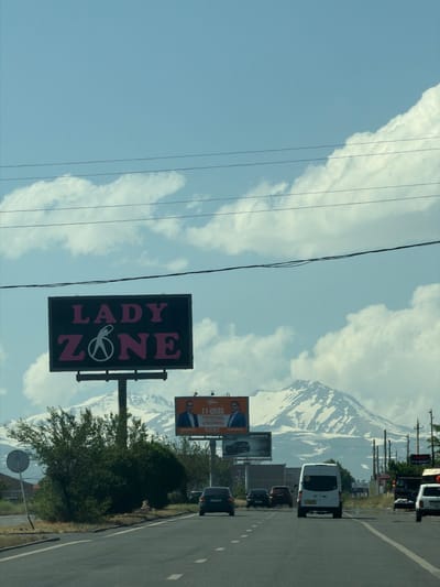A wide-angle, vertical outdoor shot captures a sunny day in Proshyan, Armenia, with snow-capped mountains in the background. The foreground shows a paved road with multiple lanes, bordered by a shoulder and a grassy area. Several cars, including a white van, are driving on the road. A large billboard displaying "LADY ZONE" in pink letters is prominent in the mid-ground, featuring a white graphic of a woman. To the right of this billboard, another smaller billboard shows two men. The sky is blue with scattered white clouds, and power lines are visible overhead. Green trees and bushes are present along the roadside. The overall scene suggests a suburban or highway setting.