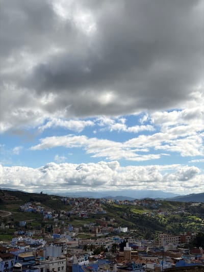 The image presents a high-angle panoramic view of the city of Chefchaouen, Morocco, situated amidst green, undulating hills. The sky occupies the upper two-thirds of the frame, featuring dark grey, overcast clouds in the top portion that transition to a bright blue sky with numerous white, puffy cumulus clouds in the mid-section.
The city's architecture, characteristic of Chefchaouen, consists of densely packed buildings predominantly painted in various shades of blue and white, with reddish-brown or grey rooftops. These structures are built into and across the hillsides, with green vegetation, including trees and shrubs, interspersed among them and covering the less developed slopes. Winding roads are visible traversing the greener hills. In the lower right quadrant, a brown mosque minaret with a green-tiled top stands out among the buildings. Distant mountain ranges appear hazy blue in the background. No individuals or discernible actions are present within the wide, static landscape view. The scene is illuminated by sunlight, indicating a partially clear day despite the significant cloud cover.