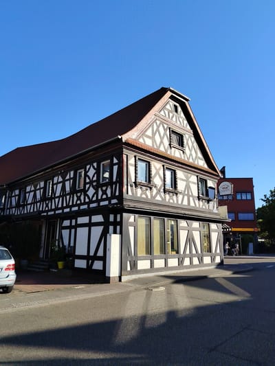 The image captures a sunny day in Achern, Germany, showcasing a traditional half-timbered building with a distinctive black and white facade. The building, which appears to house a restaurant named "China Thai Wok," has a red tiled roof and several windows, some with decorative shutters. To the right of the half-timbered building, a modern red building is visible, with shops on the ground floor. Two people are walking on the sidewalk in front of the red building. A silver car is parked on the left side of the image, near the entrance to the half-timbered building. The scene is set on a paved street, with shadows cast by the buildings and trees on the sunny asphalt. The sky is clear and blue.