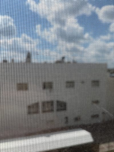 The image shows a view through a window screen at a building with a flat roof and a light-colored facade. The facade has several dark rectangular shapes, possibly windows or ventilation openings, arranged in rows. Above the building, the sky is visible with patches of blue and white clouds. The lower portion of the image shows what appears to be a reflective surface, possibly a balcony or another building feature, partially obscured by the screen. The scene is an outdoor urban environment, specifically in the city of Mirnoye, Ukraine.