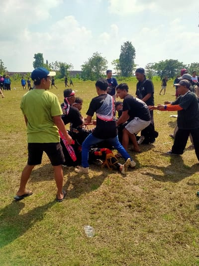 The image depicts an outdoor scene on a dry grass field in Celep, Indonesia, under a bright, partly cloudy sky, with strong sunlight casting distinct shadows. In the center-right, approximately ten adult males are actively engaged with a long, dark, cylindrical object, partially resting on the ground. This object features red and gold/yellow fringed or tasseled decorations on its visible end. Participants, dressed in a mix of casual attire including black t-shirts (one with "Seni" and a graphic print on the back), blue jeans, and shorts, are physically pulling and pushing the object. One participant wears a red and white patterned headband. To the left foreground, a male in a light green short-sleeved shirt, dark shorts, a blue baseball cap, and flip-flops stands observing the central activity, holding a red and white patterned cloth in his right hand. A crumpled, clear plastic bottle lies on the grass in the lower center foreground. The background shows numerous additional individuals scattered across the expansive field, with trees and distant structures visible on the horizon.
