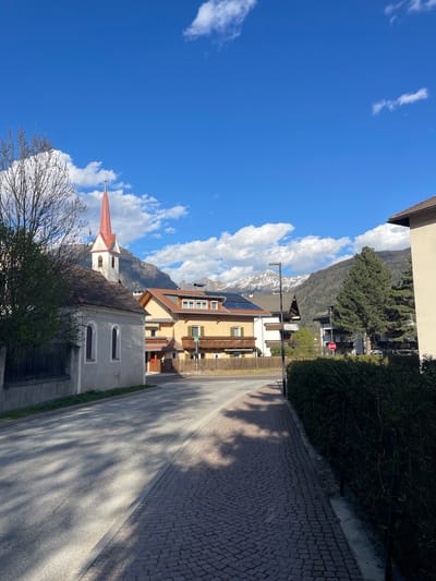 The image depicts a street scene in Sterzing-Vipiteno, Italy. A church with a distinctive red spire is visible on the left, partially obscured by bare tree branches. Adjacent to the church is a taller building with solar panels on the roof. In the background, snow-capped mountains rise behind the buildings. The sky is a vibrant blue with scattered clouds. A paved road extends into the distance, flanked by a stone-paved sidewalk. There is a stop sign near the yellow building. There are no people visible in the scene.