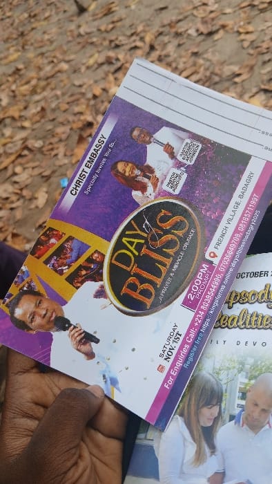 A dark-skinned hand holds a Christ Embassy flyer titled "DAY of BLISS - A PRAYER & MIRACLE CRUSADE." The flyer, predominantly purple and white, features images of Pastor Chris Oyakhilome, Pastor Bamahch Ahyntoe Johnson, and Pastor Martha Agadin, all holding microphones. It announces the event for "SATURDAY NOV. 1ST" at "2:00PM PROMPT" at "FRENCH VILLAGE, BADAGRY." Contact phone numbers and a registration URL are printed vertically on the right side. Partially visible beneath the main flyer is another printed document showing a Caucasian couple. The background consists of an outdoor ground surface thickly covered with dry, reddish-brown fallen leaves and exposed earth. The specific location within Awjaw, Nigeria, where this photograph was taken, is not discernible from the image data; the flyer advertises an event in Badagry, Nigeria.