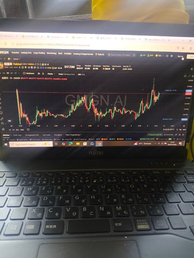 This is a close-up, eye-level shot of an open laptop displaying a cryptocurrency trading website. The website shows a real-time chart with candlestick patterns and various trading metrics such as price, volume, and time. The laptop is a Fujitsu model, and the keyboard has Japanese characters on the keys. The screen resolution appears to be standard. There are no people visible in the image. The background is blurred and appears to be a domestic or office environment, with hints of yellow and possibly furniture. The timestamp on the screen indicates "09/07/2026," and the location is given as "33°C Berawan," which translates to "33°C Cloudy." While the prompt asks for the location in Kebonbaru, Indonesia, there is no information within the image itself that corroborates this specific geographical location; the visible location data relates to weather and date.