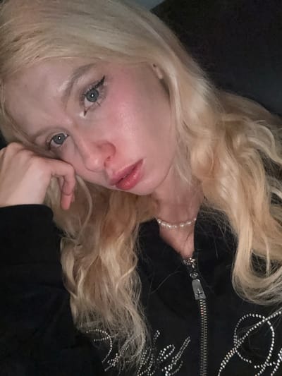 A close-up image depicts an individual with fair skin, long, wavy blonde hair, and blue eyes, situated in Namyangju-si, South Korea. The subject's left hand rests against their left cheek, supporting their head, while their gaze is directed towards the camera. Eyeliner is visible on the upper eyelids. They are wearing a black zippered top with silver studded embellishments forming a design across the chest, and a pearl choker necklace around their neck. The zipper features a silver star-shaped pull. The background is dark and indistinct.