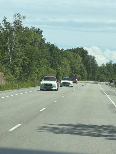 The image shows a highway with several cars. The road is paved and has white lane markings. On the left side of the road, there is a dense forest with tall trees. The sky is cloudy. The cars in the image appear to be prototypes, as their front ends are covered with camouflage material. The leading car is red, followed by a white car, and then a red SUV. Further down the road, another car is visible. The scene is set in a rural or suburban area, possibly on the outskirts of a city. The location is identified as Aur, Russia.
