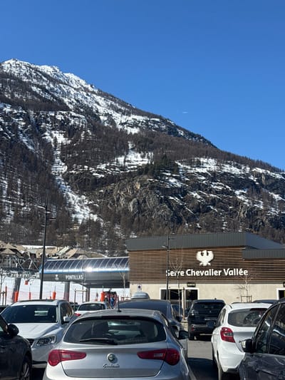 This image depicts a sunny winter or early spring scene in La Salle-les-Alpes, France, specifically at the Serre Chevalier Vallée ski resort near the Pontillas lift. In the foreground, a parking area is filled with various parked vehicles, including a silver Alfa Romeo Giulietta hatchbacks positioned centrally, a white Volvo SUV on the left, and a white hatchback on the right.
In the midground, a modern building with a facade featuring horizontal wooden slats and dark panels is visible on the right. An illuminated sign on this building reads "Serre Chevalier Vallée" below a white eagle logo, with "SORTIE" (Exit) indicated below the main text. To the left of this building, a large, covered ski lift station, identified by the label "PONTILLAS," spans across the frame. The transparent cover of the lift station reflects bright sunlight. Snow-covered ground with sparse red safety netting is visible between the parked cars and the lift station.
The background is dominated by large, snow-dusted mountains with exposed rock faces and numerous dark, slender trees, indicative of bare deciduous trees or larches in winter. The sky above is a clear, bright blue with a faint, thin contrail visible in the upper right quadrant. No identifiable individuals or specific actions are discernible in the immediate foreground, with only distant figures on the ski slopes.