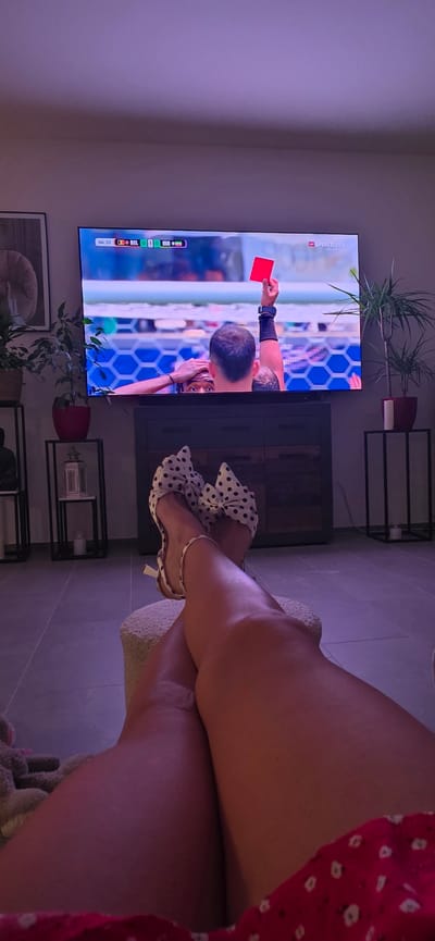 The image shows a television screen displaying a soccer match where a player is being shown a red card. The score displayed on the screen is 0-0. The television is mounted above a wooden cabinet. In the foreground, a person's legs are visible, crossed, and they are wearing polka-dot heels with large bows. The setting appears to be a living room, with a floor lamp and houseplants visible in the background. The overall lighting is dim with a purple hue. This scene is located in Lieli, Switzerland.