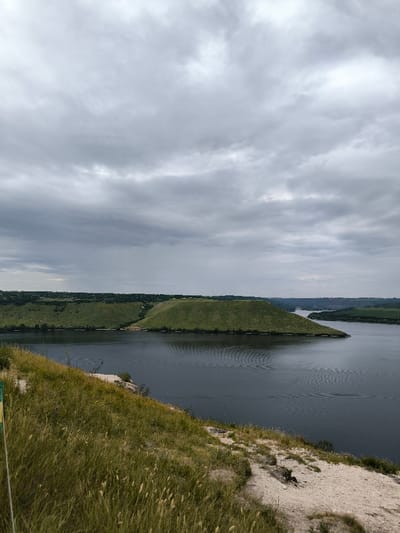 The image presents a panoramic view of a natural landscape in Horaivka, Ukraine, under an overcast sky. In the immediate foreground, a steep embankment covered in green and yellowed grasses, interspersed with areas of exposed light-colored soil or rock, descends towards a body of water. A vertical, partially obscured yellow-green object is visible on the extreme left edge of the frame. The midground features a wide expanse of dark blue-grey water, exhibiting numerous ripple patterns across its surface, indicative of water movement. Centrally positioned within the water is a distinct, verdant, flat-topped landmass, appearing as an island or a peninsula. The background consists of rolling green hills and landforms along the opposite bank of the water, stretching into the distant horizon where the water body narrows. The sky is entirely covered by layered grey clouds, suggesting overcast conditions. No human or animal subjects, explicit structures, or active interactions are discernible within the frame.