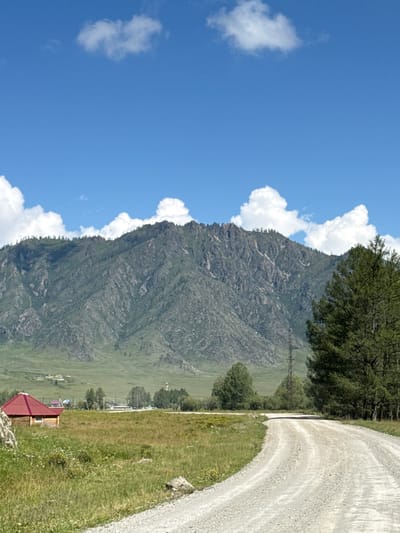 The image depicts a rural mountainous landscape in Bichiktu-Boom, Russia. The foreground features a gravel road curving to the right, bordered by a grassy field with scattered rocks and wildflowers. To the left of the road, a small wooden house with a red roof is visible. In the middle ground, there are scattered trees and a power pylon, with a small cluster of buildings further in the distance. The background is dominated by a large, rugged mountain range covered in green vegetation and rocky outcrops, under a bright blue sky with scattered white clouds. The scene appears peaceful and natural, with no people or animals present.
