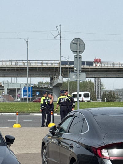 Two traffic police officers in dark uniforms and neon yellow trim stand next to a dark grey vehicle. Lettering reading "ДПС" is visible on the officers' uniforms. The setting appears to be the side of a highway or major road with white and yellow traffic markers. In the background are road signs in Russian and a white van. The presence of the Russian road signs and "ДПС" labeling on the uniform strongly suggests the location is Bolshiye Kabany, Russia. Infrastructure such as a raised platform or overpass is also visible.