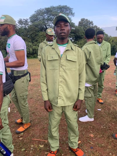 The image depicts a group of at least five young adult males standing outdoors in Kubi A, Nigeria. The central figure, a Black male, stands facing the camera, wearing a light olive-green long-sleeved collared shirt with two buttoned chest pockets, matching cargo trousers, and orange shoes with black soles. A white undershirt featuring green and white stripes is visible at his neck, and he wears a light olive-green cap with a prominent green and white emblem. To his immediate left, a partially visible Black male wears a white short-sleeved t-shirt with "NYSC" branding, light olive-green cargo trousers, orange and black shoes, a black fanny pack across his waist, and a similar light olive-green cap. Several other individuals, also Black males, are present in the background, dressed in full light olive-green uniforms consistent with the National Youth Service Corps (NYSC), standing in various orientations. The ground consists of reddish-brown soil mixed with sparse, dry grass and scattered small debris. The background features dense green trees and foliage, suggesting an open, natural environment under a pale sky, indicating a daytime setting.