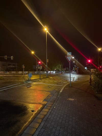 The image depicts an intersection in Hanau, Germany, at night. The street appears wet, reflecting the bright lights of street lamps and traffic signals, creating streaks of light across the scene. A traffic light displays a red signal. Various traffic signs are visible, including a yield sign with a bicycle symbol and directional signs. A building with advertising posters is in the background. The ground is paved with cobblestones on the sidewalk and asphalt on the road, with white lane markings. A discarded piece of cardboard and a small red object are on the sidewalk. The overall atmosphere is dark and illuminated by artificial light.