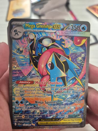 The image shows a close-up of a Mega Greninja EX Pokémon card. The card is held by a person's hand, which is partially visible on the left side of the frame. The card features a blue and pink anime-style illustration of a Pokémon, with Japanese text and English text detailing the Pokémon's abilities, HP (350), and attack power (120+). The background of the card is a colorful, abstract pattern. The surrounding environment is out of focus, but appears to be an indoor setting. There is no information in the image to suggest the location is Hagerstown, United States of America.