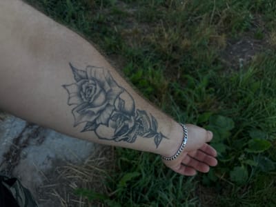 The image shows a close-up of a person's forearm with a tattoo of a rose and a skull. The person is wearing a silver bracelet on their wrist. The background is blurred and appears to be an outdoor setting with grass and some dry vegetation. The image was taken in the city of Ves', Russia.