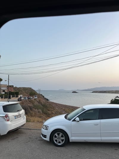 The image depicts a view from the window of a vehicle looking out towards the coast of Bolnuevo, Spain. In the foreground, two white cars are parked on an asphalt road. The car on the left is a Toyota, and the car on the right is an Audi. Beyond the road, a dry, grassy hillside slopes down towards the sea. Several other vehicles are parked along the roadside further up the hill. In the distance, the sea stretches out to meet a hazy horizon, with islands and the coastline visible. A small gazebo with a thatched roof is situated on the far coast. Power lines are strung across the sky above the scene. The sky is clear and pale blue, suggesting it is either early morning or late evening.