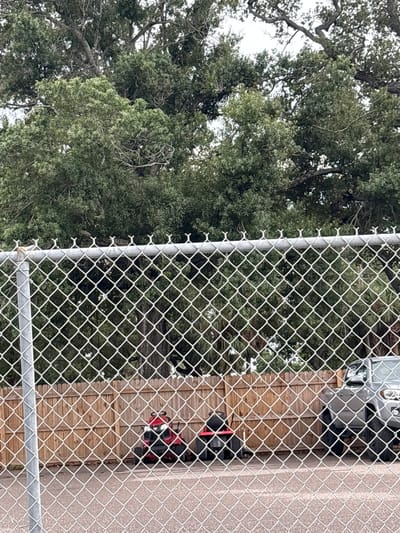 A chain-link fence runs horizontally across the foreground, obscuring a view of a yard. Behind the fence, two red riding lawnmowers are parked side-by-side in front of a light brown wooden fence. To the right of the lawnmowers, the front end of a silver SUV is visible. Lush green trees, some with visible branches, fill the background. The scene appears to be an outdoor storage area. The image was taken in Palmetto, United States of America.