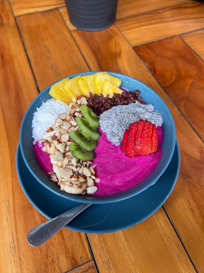 A blue bowl sits on a blue saucer atop a wooden table. The bowl contains a pink smoothie topped with chopped almonds, kiwi slices, pineapple slices, chia seeds, shredded coconut, and a strawberry. A silver spoon lies on the saucer, partially off-screen to the left. The location is Santa Fe, Costa Rica, specifically at coordinates 9.642, -85.165. The background is out of focus, but a dark pot can be seen off-screen to the top left.