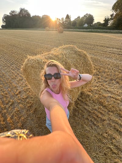 In a rural field in Trouville-Alliquerville, France, a blonde woman with blonde hair, wearing sunglasses, a pink sleeveless top, and blue jeans, is taking a selfie. She is sticking her tongue out and making a peace sign with her right hand. She has a bracelet on her left wrist. In the background, two hay bales are visible. The field is covered in straw. The sun is setting in the background, casting a warm glow over the scene.