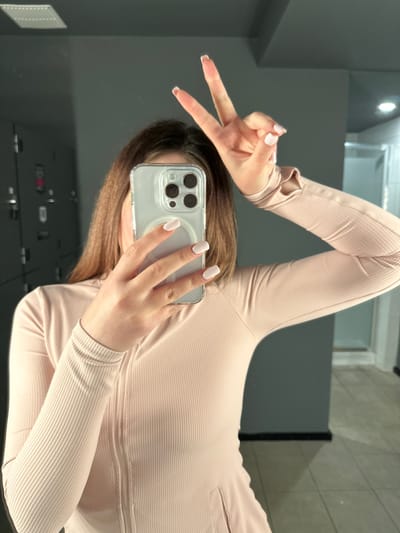In a locker room environment, a person with brown hair is holding a smartphone in their right hand, taking a selfie in a mirror. Their left hand is raised, with the index and middle fingers extended in a peace sign. The person is wearing a light pink, ribbed long-sleeved shirt with thumbholes. The background shows grey lockers and a tiled floor. The scene does not contain any identifiable landmarks or elements that would place it in Carlow, Ireland.