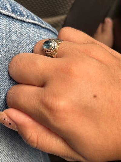 The image shows a close-up of a person's hand wearing a class ring. The ring is gold-toned with a light blue stone and inscriptions around the band. The hand is resting on blue denim fabric. In the background, a blurred foot is visible, suggesting the person might be sitting. There is no indication of the location being Conuco Viejo, Venezuela. The image is a still photograph, and no actions or interactions are depicted. The environment appears to be indoors or a shaded outdoor area.