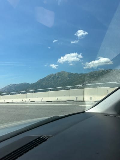 This is a photograph taken from inside a vehicle, looking out towards a mountain landscape. The view is framed by the car's interior, with the dashboard visible in the foreground and the windshield occupying the upper portion of the image. The exterior scene consists of a bright blue sky with scattered white clouds, and a range of rugged, green-covered mountains in the distance. In the middle ground, a concrete highway barrier separates the road from the mountains. The road itself is visible in the lower part of the image, with white lane markings. The scene depicts a journey through a mountainous region, likely on a highway, possibly near Bolesestra, Montenegro, given the context. There are no people or specific actions depicted, only the environment as seen from a moving vehicle.