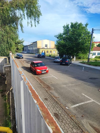 The image depicts an outdoor scene in a parking lot next to a road, likely in an industrial or commercial area, given the presence of buildings with administrative or factory-like facades. A prominent red car, a Škoda Fabia, is parked on a paved area beside a sidewalk made of interlocking stones. Other vehicles, including a black Volkswagen Golf, a dark blue car, a silver Volkswagen Polo, and a white car, are also visible in the parking lot and on the road.
The setting includes mature trees providing shade, one with drooping branches on the left and a large, full tree on the right. Buildings in the background have a functional appearance, with one notably painted in yellow and cream with balconies. A utility pole with wires is visible to the right.
The time of day appears to be late afternoon or early evening, as suggested by the warm sunlight casting shadows. The sky is blue with scattered clouds, indicating fair weather. A rusty railing runs along the left side of the foreground, suggesting the viewpoint is from an elevated position, possibly a bridge or walkway.
Visible text includes license plates on the cars. The red Škoda Fabia has the plate "2TS 5280". The black Volkswagen Golf has the plate "2UD 5518", and the silver Volkswagen Polo has the plate "6TA 0771". The overall impression is a mundane, everyday scene of parked vehicles and buildings under a pleasant sky.