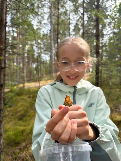A young girl with blonde hair and clear glasses is holding a small butterfly on her finger in a forest. She is wearing a light green jacket with a grey stripe. The background is a blur of green trees and sunlight. The girl is smiling and looking at the butterfly. A clear plastic container is visible at the bottom of the frame. The scene appears to be set in a natural environment, possibly a park or wooded area, consistent with the description of the city of Mucenieki, Latvia, which is known for its forests and natural landscapes.