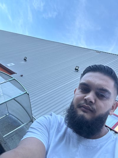 The image is a selfie taken outdoors, likely in the city of Boom, Belgium, given the context. A man with dark hair, a full beard, and a black eye is in the foreground, looking at the camera. He is wearing a white t-shirt. Behind him, a large, corrugated metal building rises steeply. The sky above is blue with wispy white clouds. Part of a translucent, arched structure, possibly a covered walkway or greenhouse, is visible on the left side of the frame. Several exterior lights are mounted on the building's wall. A white rectangular sign with the number "18" is also visible on the building's facade. The overall impression is of an industrial or commercial area.