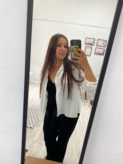 A young woman is taking a mirror selfie in a room. She is wearing a black slip dress and an open white button-down shirt over it. She has long, brown hair and is wearing a necklace. She is holding a smartphone in her right hand and has a ring on her left ring finger. The room has white walls and a light wood floor. In the background, there are several framed certificates on the wall. A rug with a zig-zag pattern is visible on the floor. The scene appears to be located in a home or a business establishment. The image was taken in Akyar, Russia.