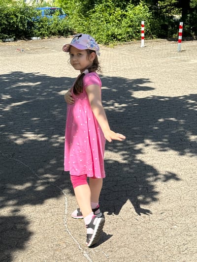 This is a vertical, eye-level, medium shot of a young girl in Wolfsburg, Germany. She is lying on her side on an asphalt surface, with her head propped up on her hand. The girl is wearing a pink dress with white polka dots, pink leggings, a pink and white patterned cap with Mickey Mouse on it, and black and white sneakers. Her right arm is extended, with her hand open as if to catch something. The girl has brown hair styled into two pigtails.
The setting appears to be outdoors during the day, as evidenced by the strong shadows cast by trees on the asphalt. The sun is bright, suggesting a clear day. To the left of the girl, there is a lush green hedge, and behind it, a blue van is partially visible. There are also two striped bollards, one red and white and the other white, on the left side of the image. The asphalt surface has some white markings on it, resembling chalk lines. The overall mood of the image is lighthearted and playful, with the girl's pose and expression suggesting she is enjoying herself. No visible text is present in the image.