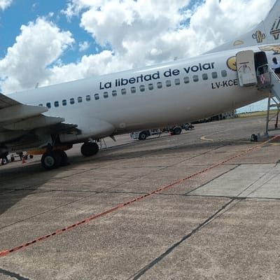 A side view of a white and yellow airplane, bearing the words "La libertad de volar" and the registration "LV-KCE", sits on a concrete tarmac under a partly cloudy sky. A boarding bridge is connected to the aircraft. Ground support equipment and safety barriers are visible near the plane's landing gear, indicating it is stationary and likely either preparing for departure or recently arrived.