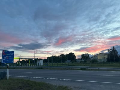 The image depicts a road scene at dusk in Laagri alevik, Estonia. A blue highway signpost on the left indicates directions to Pärnu, Haapsalu, Paldiski, Saku, Harku, and Laagri Keskus. Beyond the signpost, a Neste gas station with bright green lights is visible. The sky is filled with dramatic clouds, tinged with pink and orange hues from the setting sun. Trees and buildings line the background, with a grey road in the foreground. The overall impression is a serene evening landscape.