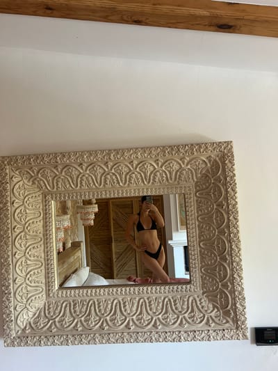 In the image, a woman in a black bikini is reflected in a large, ornate, cream-colored mirror. She is standing with one hand on her hip and the other holding a phone, appearing to take a selfie. The mirror is elaborately carved with decorative patterns. The reflection shows a bedroom setting behind her, with a bed that has white pillows and a coral-colored throw. There are wooden doors and a chandelier with shell-like elements visible in the background of the reflection. The overall lighting suggests an indoor daytime setting. The room appears to be part of a residence. The image is a photograph taken from a slightly upward angle, focusing on the mirror and its reflection.
