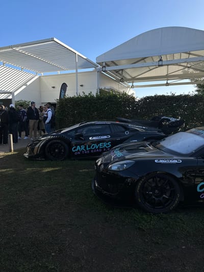 The image depicts an outdoor automotive event in Le Mans, France, under a clear blue sky. In the foreground, two heavily modified black sports cars are displayed on a patch of green grass.
The car in the mid-ground, a black Lamborghini Huracán-style race car, features extensive aerodynamic modifications including wide fenders, exposed slick tires with black multi-spoke wheels, and a prominent rear wing. White and teal lettering on its side reads "CAR LOVERS ON THE ROAD 3.0," with additional branding visible, including "LAMBORGHINI PARIS," "SCHUMACHER GP," and "E-MOTION." Gold/bronze striped accents are present on the lower side panel.
Partially visible in the immediate foreground, a second black sports car, possibly an Alpine A110 variant, exhibits similar race-prepared styling. It features black multi-spoke wheels with "PIRELLI P ZERO" text visible on the tires and "E-MOTION" branding near the headlights.
Behind the vehicles, a group of approximately 10-15 individuals, predominantly male and casually dressed, are gathered on the grass. One person in the center-left is holding a phone to their ear. They appear to be spectators or event attendees.
The background features two temporary structures. To the left, a white building with a flat, slatted white metal canopy extends outwards, casting distinct shadows. To the right, a large white arched tent-like structure is visible. A dense, dark green hedge separates the parked cars from these structures. A dark banner with light-colored branding is partially visible near the building on the left. The ground underfoot is primarily grass, with some areas appearing worn.