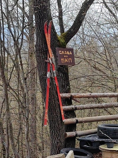 An outdoor scene shows a tree with a pair of vintage red skis leaning against its trunk. Attached to the tree above the skis is a brown sign with text in Cyrillic script. A section of a wooden fence constructed from horizontal logs and dark rubber tires rests below and slightly to the right of the tree. The background consists of a forest environment with numerous trees, many of which are bare branched and a cloudy sky. The scene is located in the city of Dushevo, Bulgaria.