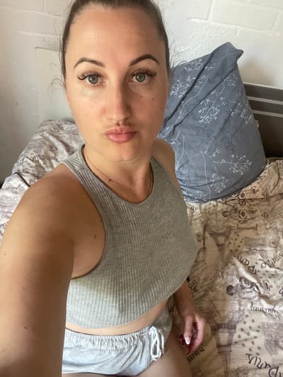 In Zarichne, Ukraine, a woman with dark hair pulled back is positioned on a bed. She is wearing a grey ribbed tank top and grey shorts. Her eyes are open, and her lips are slightly puckered. The bed is covered with patterned sheets and a blue pillow with a white leaf design. The background shows a white brick wall.