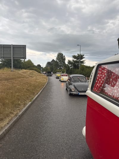 The image shows a procession of vintage Volkswagen cars driving on a wet asphalt road. In the foreground, a red and white vehicle, likely another vintage Volkswagen, is partially visible from the side. Several Volkswagen Beetles are visible in the procession, with one dark grey Beetle in the middle ground displaying the license plate "ASS 02". Yellow and white Beetles are also part of the line. The road is bordered on the left by a grassy embankment and on the right by trees. The sky is overcast and grey. The scene appears to be on a highway or a main road, possibly near the city of Falkenberg, Sweden, as suggested by the presence of Swedish flags on some of the vehicles and the overall landscape. The cars are driving in the same direction, indicating a parade or a group outing.