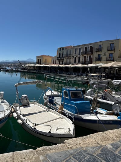 The image depicts the Venetian harbor in Rethymno, Greece. In the foreground, several small boats are moored. The closest boat on the right has a white hull with a blue stripe and appears to be a fishing boat, as indicated by the netting and ropes. It has the registration "ΖΕΦΥΡΟΣ ΛΡ. 1200" on its side. Next to it, to the left, is another white boat with a light-colored canopy. The leftmost boat in the immediate foreground is partially visible, showing its white hull.
In the mid-ground, more boats are docked, some with their covers on. Beyond the boats, a promenade is lined with buildings featuring arched windows and balconies. Several establishments with outdoor seating and umbrellas suggest a waterfront dining or cafe area. The architecture of the buildings is characteristic of Venetian influence, with pastel-colored facades and terracotta roofs. A prominent sign on one building reads "DEA DEL PORTO."
The background reveals a calm body of water reflecting the sky and buildings. In the distance, hills and mountains are visible under a clear, bright blue sky, suggesting a sunny day. The overall scene conveys a peaceful and picturesque maritime atmosphere in the historic city of Rethymno.