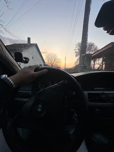 The scene depicts an interior view from a vehicle, looking through the windshield towards a street or rural setting in Jitnitsa, Bulgaria, during either sunrise or sunset.

**People:** A single individual's right hand is visible in the lower left quadrant, resting on the steering wheel. This person is wearing a silver-colored watch with a block-link metal band on their wrist.

**Subjects/Objects (Interior):** The interior of a dark-colored BMW vehicle is visible. The prominent object is a black leather steering wheel with the BMW emblem at its center. The instrument cluster, partially obscured by the steering wheel, shows a tachometer/speedometer dial. Air vents are visible on the dashboard to the right. A set of keys hangs from the ignition or steering column. A black mobile phone mount with a suction cup base is affixed to the windshield on the upper left.

**Subjects/Objects (Exterior/Environment):** The sky displays a gradient of colors, transitioning from light blue at the top to warm orange and pink hues near the horizon, indicative of twilight. Multiple overhead utility lines, likely power lines, crisscross the upper sky. On the left, a light-colored, single-story house with a reddish-brown tiled roof and a chimney is visible. To the right, a tall, dark utility pole stands prominently. Beyond the pole, a tree with bare branches or early pinkish blossoms is seen. Further to the right, a rustic wooden structure with an open-sided, gabled roof is partially visible. The ground beyond the vehicle is not distinctly visible but suggests a road or unpaved area.
