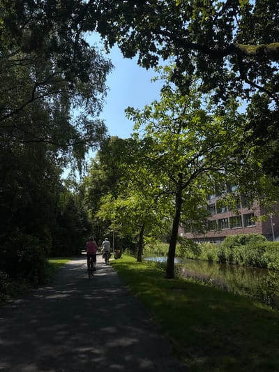 The image depicts a paved pathway in Gronau, Germany, bordered by lush green trees and vegetation. Two individuals are seen cycling away from the camera on the pathway. The path runs alongside a tranquil body of water, reflecting the surrounding trees and a modern brick building visible in the background. The scene is bathed in sunlight, with dappled shadows cast across the pathway from the overhanging trees.