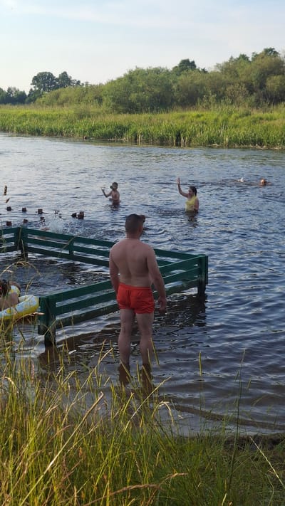 In an outdoor scene, likely a river or lake in Навасвержанскі сельскі Савет, Belarus, several individuals are enjoying a summer day. A man stands in the foreground, facing away from the camera, wearing orange swim trunks. He is partially submerged in the water, with tall grass and reeds at the edge of the water obscuring his lower legs. In the middle ground, a green wooden structure, possibly a jetty or a part of a swimming area, extends into the water. Further back, several other people are visible in the water. One person, wearing a yellow top, is raising their arm. Two children are also in the water; one is splashing, and another appears to be standing. The background is dominated by lush green trees and vegetation, suggesting a rural or natural setting. The sky is clear and blue.