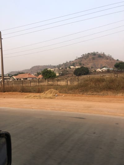 A roadside view features utility poles and lines against a hazy sky. Adjacent is a scrubby verge and a road surface with indistinct markings. Beyond, a concrete wall lines the edge of residential properties nestled at the foot of a brown, arid hill dotted with trees and some buildings. The landscape appears dry, with a muted color palette dominated by browns, greys, and a hazy blue.