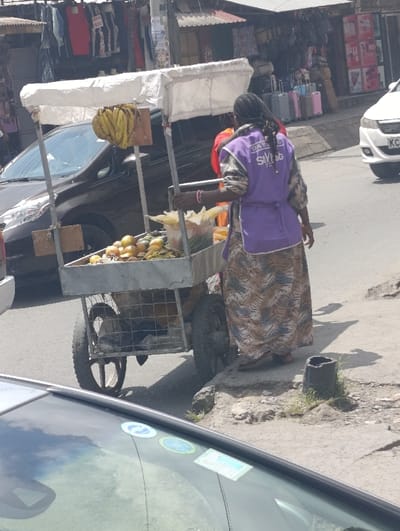 The image displays a daylight street scene in Matungulu west ward, Kenya. A female street vendor, viewed from behind, stands adjacent to a three-wheeled metal pushcart. She wears a long, patterned skirt, a light-colored long-sleeved top, and a purple vest or apron with "DARLING STYLIST" printed in white. Her dark hair is styled in braids or dreadlocks.
The vendor's cart features a white fabric canopy. A bunch of yellow bananas hangs from the front of the canopy frame. The cart's main display area holds various fruits, including yellow mangoes or oranges, some green produce, and multiple clear plastic bags containing husked corn. Below the main compartment, a metal cage structure contains dark, indistinct objects.
The vendor is positioned on an elevated, uneven concrete or dirt pavement area bordering an asphalt road. A broken concrete pipe segment lies on the pavement near her feet. To the left of the cart, a dark-colored SUV or hatchback is partially visible on the road. To the right, a white car with a Kenyan license plate (starting with "KC") is also on the road.
In the background, behind the vehicles and vendor, are several market stalls or shops. The left background shows hanging clothing items for sale, with some price tags visible (e.g., "800"). The right background features stalls displaying luggage, including pink, blue, and silver suitcases, among other goods. The immediate foreground shows the upper part of a car's windshield and roof, with several stickers affixed to the windshield. Shadows indicate a bright, sunny environment.