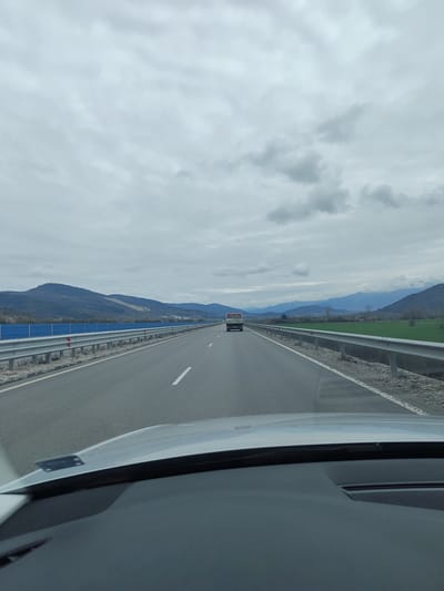 The image shows a view from inside a car driving on a highway near Novachene, Bulgaria. The road is straight and stretches into the distance, flanked by metal guardrails on both sides. A truck is visible ahead, in the same lane. Mountains are visible in the background, partially obscured by a cloudy sky. The hood of the car is visible at the bottom of the frame. A blue fence is visible on the left, along the side of the road. Green fields are seen on the right side of the road.