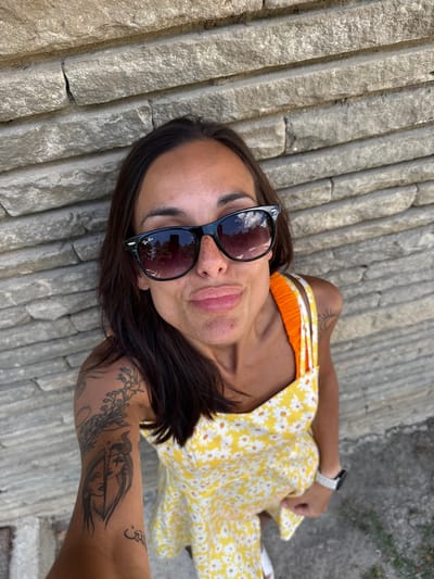 A woman stands against a stone wall in Grivitsa, Bulgaria. She is wearing a yellow dress with white flowers and orange straps, black sunglasses, and a smartwatch. Her arm is tattooed with several designs, including a face with horns and a branch. The image is taken from a high angle, looking down at the woman. The wall is made of rough-hewn gray stones.