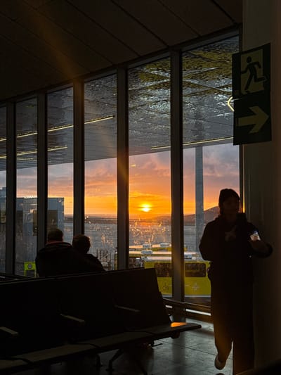 The scene is an interior view within an airport terminal located in Granadilla de Abona, Spain, at sunset. A panoramic glass window wall dominates the central and left frame, showcasing a vibrant orange sunset with a prominent bright orange sun low on the horizon. The exterior view includes what appears to be a body of water or wet ground reflecting the sky, with distant land formations and several yellow ground vehicles partially visible.
The interior foreground features multiple rows of unoccupied, dark-colored modular seating units, typical of an airport waiting area. Two individuals are seated with their backs to the camera on one of these units, observing the exterior view. A third individual stands partially silhouetted on the right side of the frame, facing left towards the windows. This person wears dark attire, and a light-colored object, possibly a badge or wristband, is visible on their right forearm.
Two green emergency exit signs, one depicting a running figure and another indicating a right-pointing arrow, are affixed to a structural column on the far right. The interior lighting is subdued, with reflections of overhead linear lights visible on the upper sections of the windows and the ceiling panels.