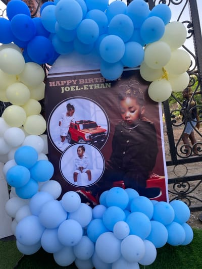 The image shows a birthday banner with a photo of a young boy, Joel-Ethan, sitting on a toy car and standing next to a red toy truck. The banner is decorated with blue and white balloons, arranged in a semi-circle around the banner. The banner is on a metal gate, and the background is a green lawn. There is a person walking out of the gate to the right of the banner. The location is Bonadikombo, Cameroon, at coordinates 4.048, 9.218.